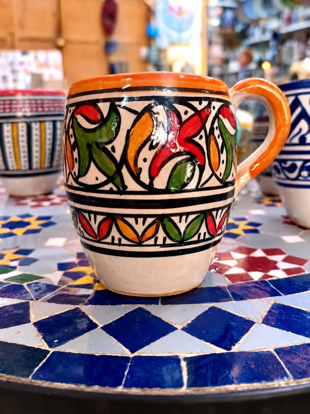Orange and multicolor hand-painted Moroccan ceramic mug displayed on traditional Moroccan mosaic tile.
